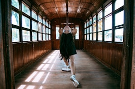 A person elegantly balances in a dance pose inside an old wooden train carriage with windows lining both sides. Sunlight streams through the windows, casting soft patterns on the wooden floor.