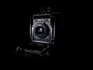 A vintage camera is prominently displayed against a dark background, showcasing its intricate mechanical details and lens at the center. The lighting highlights the contours and design of the camera, emphasizing its classic and historical appearance.