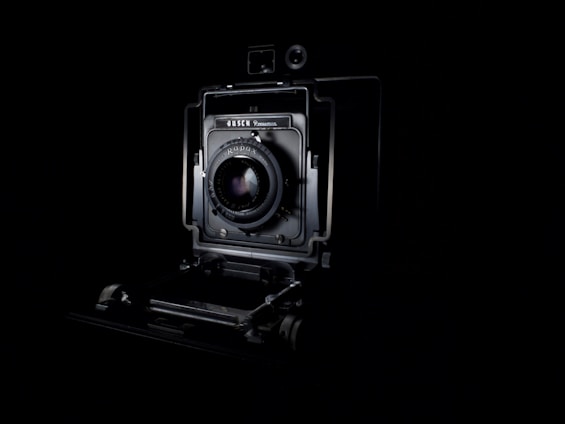 A vintage camera is prominently displayed against a dark background, showcasing its intricate mechanical details and lens at the center. The lighting highlights the contours and design of the camera, emphasizing its classic and historical appearance.