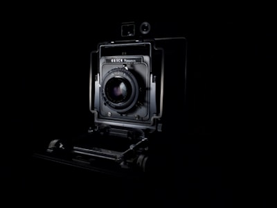A vintage camera is prominently displayed against a dark background, showcasing its intricate mechanical details and lens at the center. The lighting highlights the contours and design of the camera, emphasizing its classic and historical appearance.