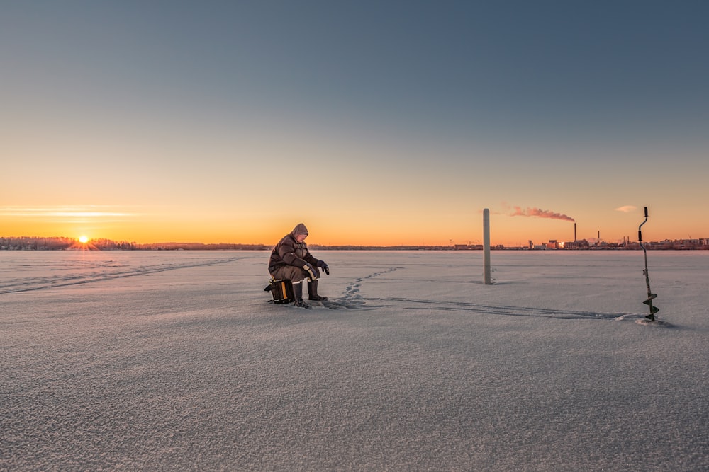 person sitting on stool on ice field near manual auger
