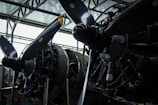 Close-up view of two large aircraft engines with propellers. The intricate details of the engines are visible, including various pipes, wires, and mechanical components. The propellers have yellow tips, and the structure appears to be housed inside an industrial building with a high ceiling.