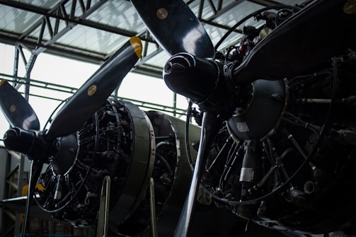 Close-up view of two large aircraft engines with propellers. The intricate details of the engines are visible, including various pipes, wires, and mechanical components. The propellers have yellow tips, and the structure appears to be housed inside an industrial building with a high ceiling.