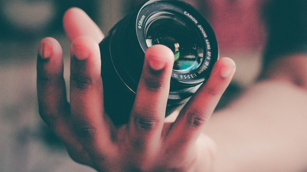 fotografia de foco raso de pessoa segurando lente de câmera DLSR preta