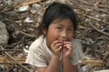 A young child with dark hair sits in an outdoor setting cluttered with debris and natural elements. The child has a contemplative expression, with hands raised near the face, and is dressed in a light-colored shirt. The background is filled with twigs, leaves, and scattered objects.