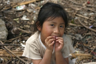 A young child with dark hair sits in an outdoor setting cluttered with debris and natural elements. The child has a contemplative expression, with hands raised near the face, and is dressed in a light-colored shirt. The background is filled with twigs, leaves, and scattered objects.