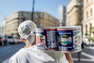 A person carrying a pack of large canned goods, featuring Italian branding, is walking through an urban street with blurred buildings and vehicles in the background. The individual is wearing a white shirt and a headscarf.