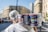 A person carrying a pack of large canned goods, featuring Italian branding, is walking through an urban street with blurred buildings and vehicles in the background. The individual is wearing a white shirt and a headscarf.