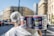 A person carrying a pack of large canned goods, featuring Italian branding, is walking through an urban street with blurred buildings and vehicles in the background. The individual is wearing a white shirt and a headscarf.