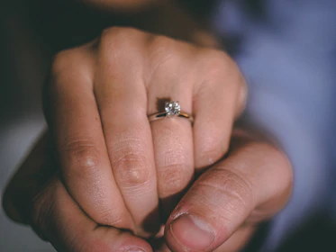 Close-up of intertwined hands showing off a sparkling engagement ring against a soft floral background.