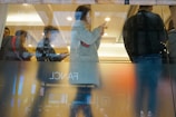 Several people are standing in a queue inside a dimly lit room, with reflections visible on the glass window. One person is prominently using a smartphone.