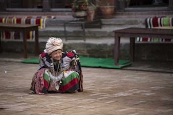 A child dressed in traditional, colorful attire sits on a brick paved ground, surrounded by various patterns and fabrics. The child wears a large headdress and holds an intricate, decorative necklace. Tables and potted plants are visible in the background.