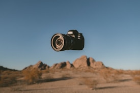 A digital camera is hovering in the air against a backdrop of a desert landscape with rocky formations in the distance. The sky is clear with a warm, golden hue.