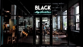 A modern caf&eacute; interior with a stylish neon sign reading 'BLACK' above an entrance. The setting is dimly lit with warm, ambient lighting and sleek, contemporary furniture. Large windows with reflections allow natural light to filter in. Patrons are seated at tables, enjoying their drinks in a cozy atmosphere.