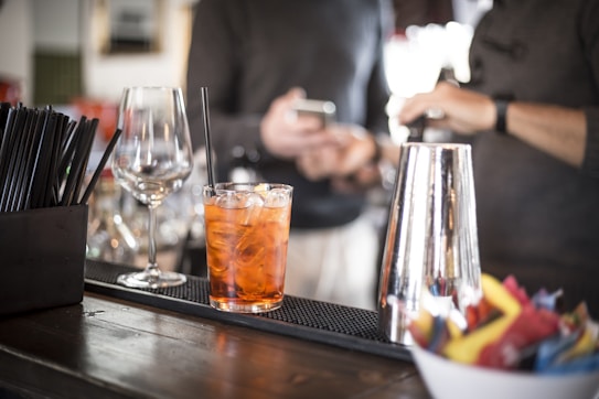 A cocktail sits on a bar counter next to a wine glass and a metal shaker. Two people in the background are blurred, one holding a phone. A container of black stirrers and colorful cocktail napkins also appear on the counter.