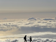 Silhouettes of two people stand atop a sea of fluffy clouds with a vast sky above. The scene suggests a sense of adventure and awe, with the figures seemingly engaged in a moment of connection or exchange.