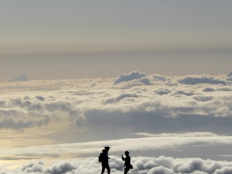 Silhouettes of two people stand atop a sea of fluffy clouds with a vast sky above. The scene suggests a sense of adventure and awe, with the figures seemingly engaged in a moment of connection or exchange.