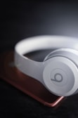 A pair of white over-ear headphones resting on a smartphone with a slightly blurred, dark background. The headphones have a prominent circular logo on the ear cup, and the smartphone has a metallic rose color.