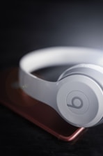 A pair of white over-ear headphones resting on a smartphone with a slightly blurred, dark background. The headphones have a prominent circular logo on the ear cup, and the smartphone has a metallic rose color.