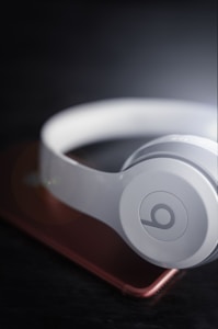 A pair of white over-ear headphones resting on a smartphone with a slightly blurred, dark background. The headphones have a prominent circular logo on the ear cup, and the smartphone has a metallic rose color.