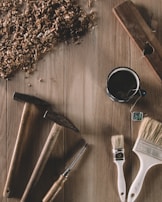 Wood shavings scatter across a wooden surface, accompanied by two hammers, a screwdriver, two paintbrushes, and a cup of tea with a tag. A plane is positioned at the top right, contributing to the woodworking atmosphere.