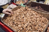 A large cardboard box filled with shelled walnuts is displayed, with a hand reaching in to pick some. Surrounding the box are other containers filled with different nuts and dried fruits, possibly part of a market stall or shop setting.