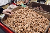 A large cardboard box filled with shelled walnuts is displayed, with a hand reaching in to pick some. Surrounding the box are other containers filled with different nuts and dried fruits, possibly part of a market stall or shop setting.