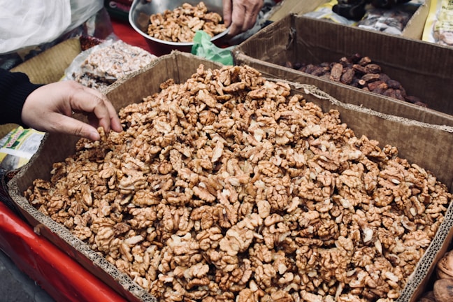 A large cardboard box filled with shelled walnuts is displayed, with a hand reaching in to pick some. Surrounding the box are other containers filled with different nuts and dried fruits, possibly part of a market stall or shop setting.