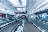 A spacious airport terminal with a modern design features metallic moving walkways and overhead screens displaying information. The architecture includes high ceilings and extensive use of glass and steel. Several travelers with luggage are visible in the distance, creating a sense of movement.