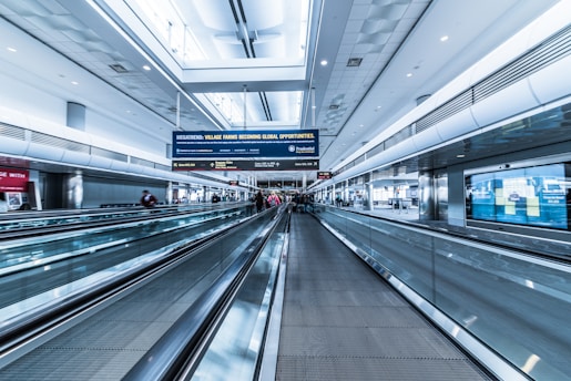 A spacious airport terminal with a modern design features metallic moving walkways and overhead screens displaying information. The architecture includes high ceilings and extensive use of glass and steel. Several travelers with luggage are visible in the distance, creating a sense of movement.
