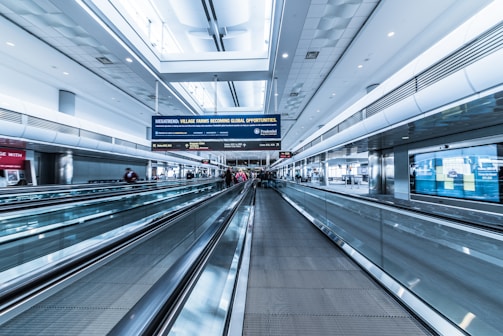 A spacious airport terminal with a modern design features metallic moving walkways and overhead screens displaying information. The architecture includes high ceilings and extensive use of glass and steel. Several travelers with luggage are visible in the distance, creating a sense of movement.