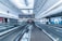 A spacious airport terminal with a modern design features metallic moving walkways and overhead screens displaying information. The architecture includes high ceilings and extensive use of glass and steel. Several travelers with luggage are visible in the distance, creating a sense of movement.