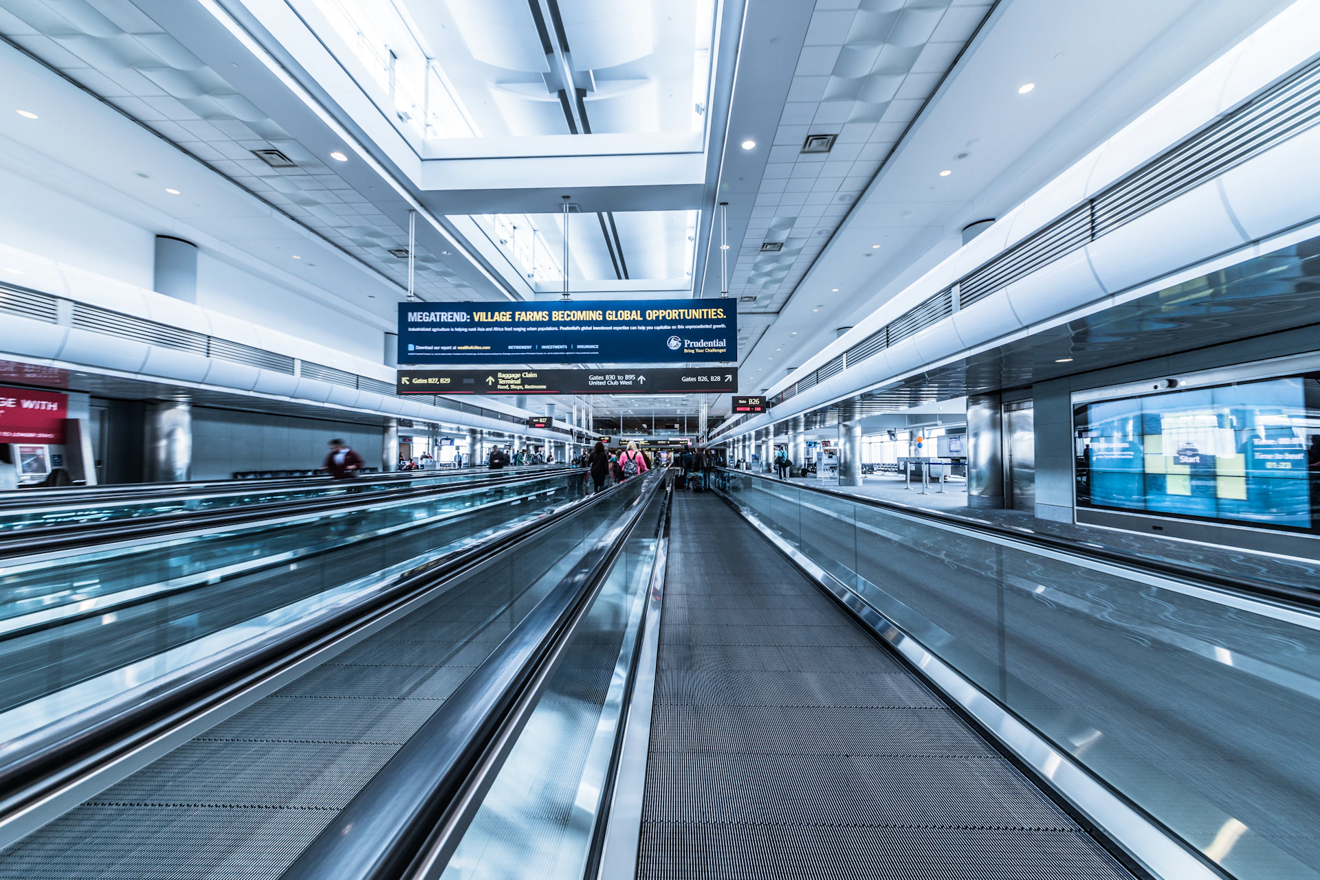 A spacious airport terminal with a modern design features metallic moving walkways and overhead screens displaying information. The architecture includes high ceilings and extensive use of glass and steel. Several travelers with luggage are visible in the distance, creating a sense of movement.