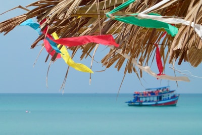 A serene beach scene with colorful fabric streamers fluttering from a thatched roof against a clear blue sky. In the background, a colorful boat is floating on the calm turquoise sea.