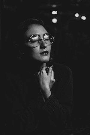 A black and white portrait of a person with short hair and glasses. The person has their eyes closed and hands clasped near their face, appearing contemplative or reflective.