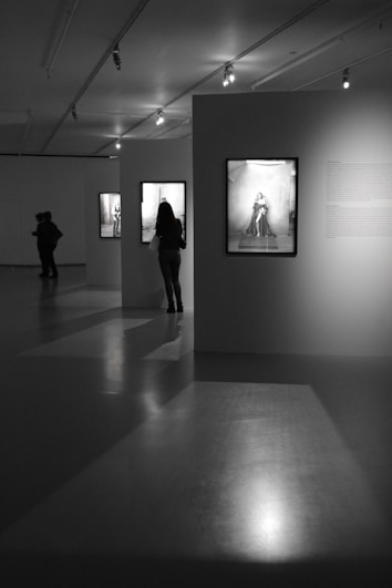 A dimly lit art gallery featuring black-and-white photographs on the walls. Silhouettes of people are observed viewing the exhibits, creating a contemplative atmosphere. The gallery space is illuminated by overhead spotlights that cast shadows on the glossy floor.