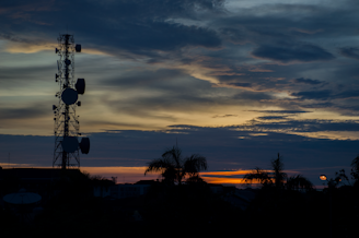 A silhouette of a communication tower stands against a striking sunset sky with layers of clouds and vibrant hues. Palm trees and rooftops are visible in the foreground, with the sun casting warm tones across the horizon.