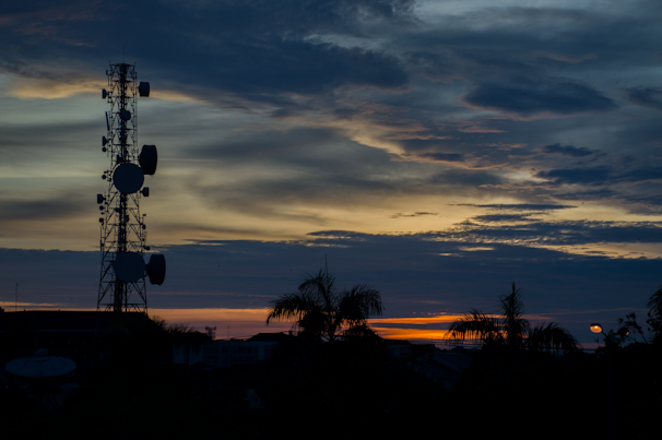 A silhouette of a communication tower stands against a striking sunset sky with layers of clouds and vibrant hues. Palm trees and rooftops are visible in the foreground, with the sun casting warm tones across the horizon.