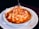 A dish of baked lasagna with a golden, cheesy topping, served in a white ceramic bowl placed on a white plate. The edges of the lasagna are slightly browned, and there are visible layers of pasta and sauce beneath the cheese. A dark-colored napkin and part of a glass stem are seen in the background.