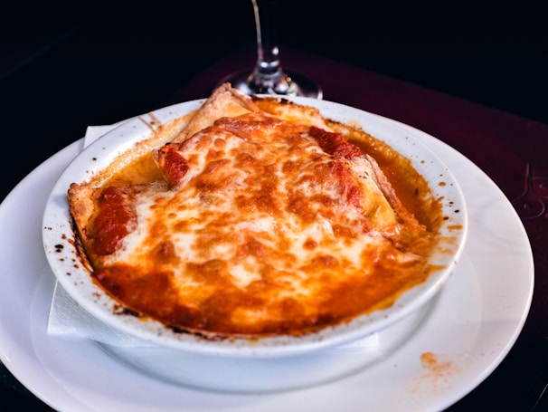 A dish of baked lasagna with a golden, cheesy topping, served in a white ceramic bowl placed on a white plate. The edges of the lasagna are slightly browned, and there are visible layers of pasta and sauce beneath the cheese. A dark-colored napkin and part of a glass stem are seen in the background.