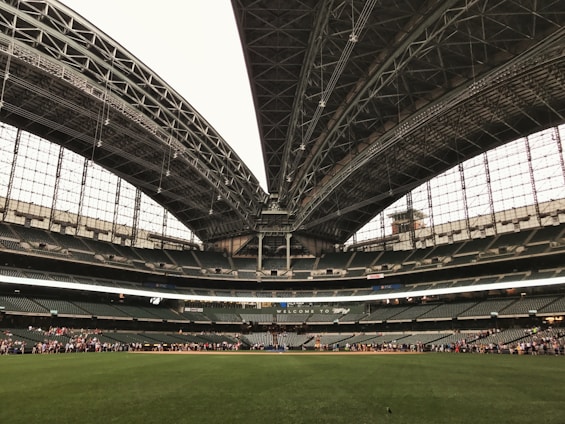 An expansive sports stadium with a large, open field of lush green grass and a partially retractable roof, showcasing a vast seating area surrounding the field. A group of people is visible along the boundary, seemingly engaged in an event or tour.