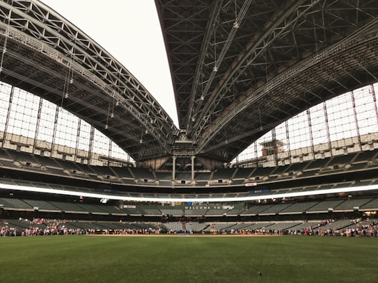 An expansive sports stadium with a large, open field of lush green grass and a partially retractable roof, showcasing a vast seating area surrounding the field. A group of people is visible along the boundary, seemingly engaged in an event or tour.