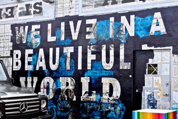 A mural featuring the phrase 'WE LIVE IN A BEAUTIFUL WORLD' painted in large, bold white letters against a dark background with blue accents. The wall is decorated with collage-like patterns and text excerpts. A black vehicle is parked partially in view on the left, with only the front visible and a colorful, striped block on the right.