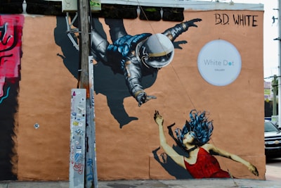 A vibrant street mural depicting an astronaut reaching towards a woman with blue hair and a red dress, set on a peach-colored wall. The artwork is partially obscured by a pole covered with stickers. The mural is signed by B.D. White and next to the White Dot Gallery sign.