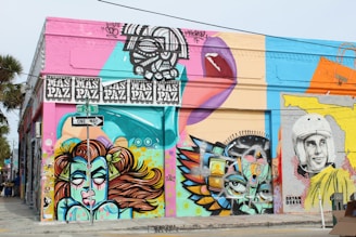 A vibrant street art mural covers the side of a building at the intersection of NW 24th Street. The artwork features a variety of colorful designs, including a stylized portrait of a woman with flowing hair, abstract geometric shapes, and a black-and-white depiction of a person wearing a vintage helmet. The wall is painted in bright hues, with bold outlines and intricate patterns. Above, there's a recurring motif with the words 'MAS PAZ'. A street sign displaying 'One Way' is visible at the junction.