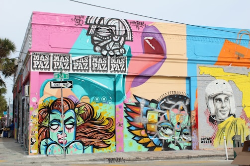 A vibrant street art mural covers the side of a building at the intersection of NW 24th Street. The artwork features a variety of colorful designs, including a stylized portrait of a woman with flowing hair, abstract geometric shapes, and a black-and-white depiction of a person wearing a vintage helmet. The wall is painted in bright hues, with bold outlines and intricate patterns. Above, there's a recurring motif with the words 'MAS PAZ'. A street sign displaying 'One Way' is visible at the junction.