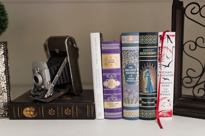 A collection of classic hardcover books with decorative covers is neatly arranged in a row. An antique folding camera is positioned beside the books. The book covers display intricate patterns and one features a vibrant red ribbon bookmark. The image conveys an atmosphere of vintage elegance and literary appreciation.