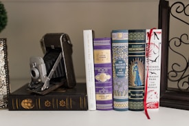 A collection of classic hardcover books with decorative covers is neatly arranged in a row. An antique folding camera is positioned beside the books. The book covers display intricate patterns and one features a vibrant red ribbon bookmark. The image conveys an atmosphere of vintage elegance and literary appreciation.
