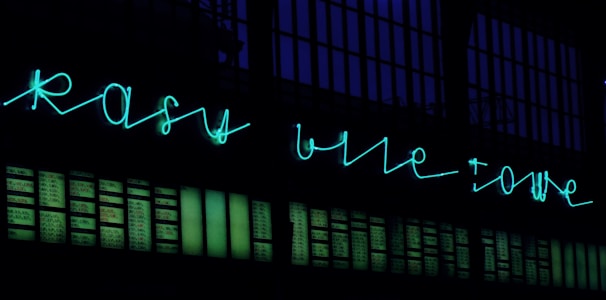 Neon sign in turquoise script displayed above a wall with numerous rectangular panels, possibly windows, illuminated against a dark background.