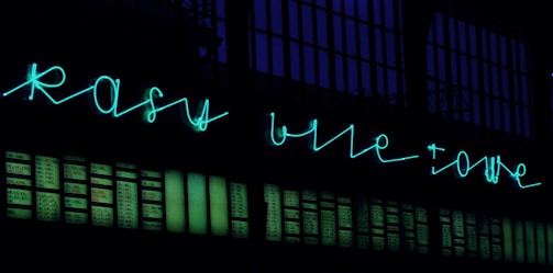Neon sign in turquoise script displayed above a wall with numerous rectangular panels, possibly windows, illuminated against a dark background.